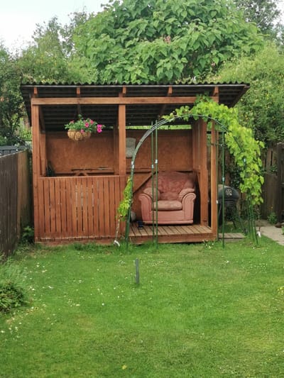 This image depicts a wooden shelter in a garden, likely in Ripon, United Kingdom, on a cloudy day. The shelter has a corrugated roof and wooden walls. Inside, a pink armchair is placed on a wooden floor. A hanging basket filled with pink flowers is suspended from the roof. To the right of the shelter, an outdoor grill is visible. A green metal archway with climbing vines is positioned in front of the shelter, extending over the grassy lawn. The lawn is green with scattered white and yellow wildflowers. A wooden fence runs along the left side of the image, and more greenery and trees are visible in the background. No people or text are present in the image. The overall impression is one of a serene, outdoor relaxation space.