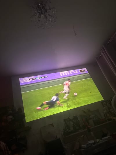 The image shows a projection of a soccer game on a wall in a room. The score displayed on the screen indicates a match between England (ENG) and Argentina (ARG) with a score of 0-0 at 31:16. Two players are visible on the grass field of the game: one in a white and red uniform with the number 10, and another in a blue and white uniform, on the ground, with the number 11. The room appears to be a living area with curtains on the right side and a chandelier visible on the ceiling. A shelf with various objects, including framed pictures and decorative items, is present on the left side of the image, below the projected screen. The lighting in the room is dim, with the projection screen being the primary light source. The location in Novyy, Russia, is not discernible from the image itself.