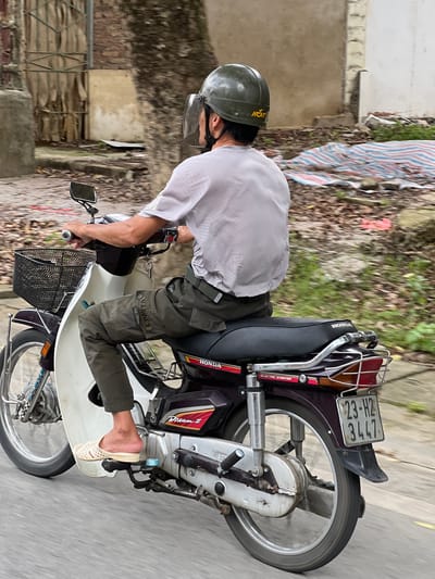 In a medium shot, a person rides a purple Honda Dream II motorcycle on a paved road in Hà Giang 2 Ward, Vietnam. The rider, viewed from the rear-right, wears a green helmet with a clear face shield and a light gray t-shirt. Their dark green cargo pants are adorned with a "MARINES" patch. They are also wearing light-colored slip-on sandals. The motorcycle has a black basket on the front and a luggage rack at the back. A license plate reading "23-H2 3447" is visible. The background shows a tree with rough bark, a weathered wall, and some foliage, with a striped tarp partially visible to the right. The scene suggests a typical street scene in Vietnam.