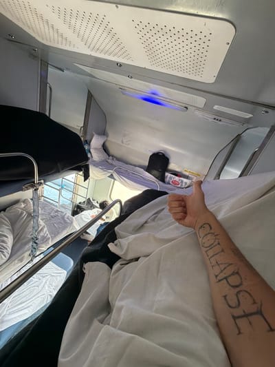 The image depicts the interior of a train carriage, likely a sleeper class compartment, in Lucknow, India. The compartment appears to be equipped with bunk beds, with white and dark bedding visible. A person's arm is prominently featured in the foreground, giving a thumbs-up gesture. This arm has a tattoo on it that reads "COLLAPSE". Other visible objects include a black backpack, some packaged goods, and what appears to be a water bottle. The ceiling of the carriage features perforated panels, possibly for ventilation or lighting. The overall impression is that of a person traveling by train and signaling their contentment with the journey.