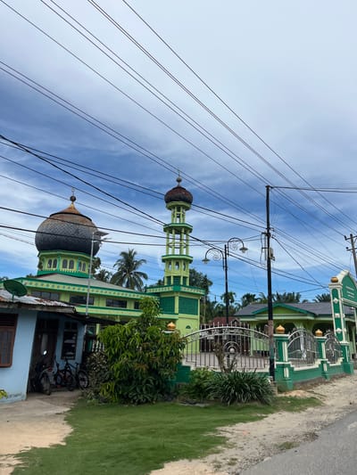 A low-angle, eye-level outdoor shot captures a mosque complex in Bukitpanjang Dua, Indonesia, under a partly cloudy sky. Prominently featured is a tall, slender green minaret with multiple levels and arched openings, topped with a golden finial. To the left of the minaret, a mosque building displays a large, weathered dome, segmented like scales and a golden spire. The mosque's facade is painted in bright green with white decorative elements and arched windows. A lower structure, possibly a house or attached building, is painted light blue with a brown wooden window and door, and a corrugated metal roof. Two bicycles and a motorcycle are parked under an overhang of this blue building. A green metal fence with decorative scrollwork encloses the mosque grounds, separating it from the street. A single lamppost with two ornate lamps stands near the fence. Lush green trees and palm trees are visible behind the mosque. The ground in front of the mosque is a mix of sand and short green grass, and a paved road is visible on the right side of the image. Numerous power lines crisscross the sky above the scene.