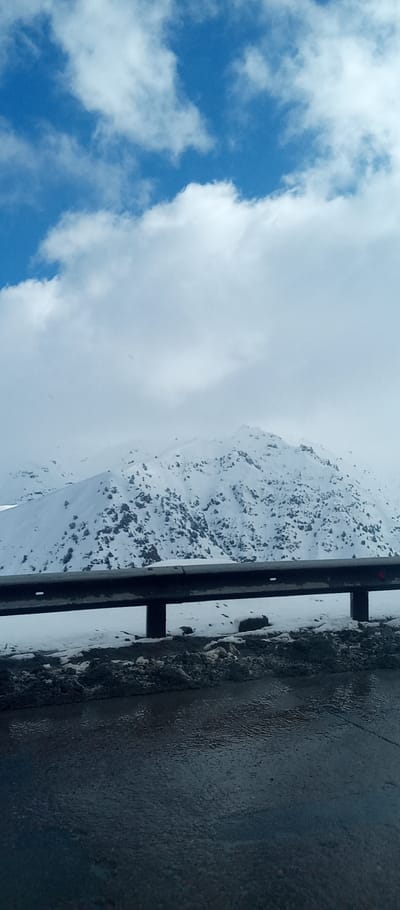 A mountain covered in snow and dotted with trees dominates the background. A guardrail stretches across the midground, with snow visible along the roadside. The foreground shows an asphalt road reflecting light. The sky is visible above the mountain and is a mix of blue and white, with scattered clouds. The scene appears to be taken from a moving vehicle.