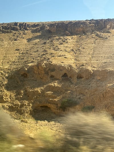 The image depicts a close-up view of a rugged, rocky cliff face, likely in a desert or arid environment, given the muted earth tones and sparse vegetation. The cliff is bathed in bright sunlight, suggesting it is daytime, possibly late morning or early afternoon. The texture of the rock is rough and uneven, with visible striations and numerous small indentations and crevices. Some larger hollows or small caves are apparent in the rock face.
To the left of the cliff, a clear, pale blue sky is visible, indicating fair weather. The right side of the image is heavily blurred with streaks of light and color, suggesting motion, possibly from a vehicle passing by or a water feature. This blur creates a dynamic contrast with the static, detailed texture of the cliff.
There are no people or man-made objects clearly identifiable in the scene. The activity is not explicitly depicted, but the context of the blurred motion suggests travel. The overall impression is one of a natural, rugged landscape under bright sunlight. The location context provided indicates Arad, Israel, which is consistent with the arid appearance of the cliff.