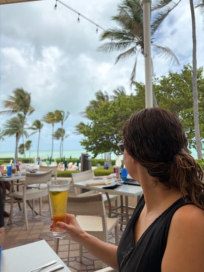 The photograph shows a woman at an outdoor dining area, likely in a resort or upscale casual restaurant, in what is determined to be the city of Key Biscayne, United States of America. The woman has dark hair, is wearing sunglasses and a black top and she is holding a glass of beer. The drink is light amber in color, with visible bubbles, and a lime wedge is visible in the glass.
The dining area features white tables and wicker chairs. In the background, palm trees are visible, along with the ocean. The presence of palm trees and the ocean indicates a tropical or subtropical setting consistent with Key Biscayne. The lighting suggests a daytime shot with slightly overcast conditions.