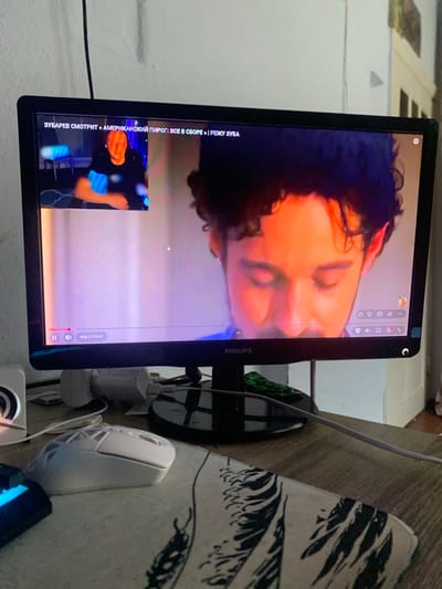 The image shows a computer monitor displaying a video. The video appears to be a movie or show, with a close-up of a person's face filling most of the screen. The person has dark, curly hair and is illuminated with warm, orange light, suggesting a dramatic or emotional scene. In the bottom left corner of the video, a smaller window shows another person, a man wearing a black hoodie, looking towards the camera. The video has a title overlay in Russian: "ЗУБАРЕВ СМОТРИТ «АМЕРИКАНСКИЙ ПИРОГ: ВСЕ В СБОРЕ » | РЕЖУ ЗУБА" which translates to "ZUBAREV WATCHES 'AMERICAN PIE: ALL TOGETHER' | CUTTING TOOTH". A timestamp at the bottom right indicates the video is at 4 minutes and 19 minutes and 41 seconds into playback. The monitor itself is a Philips brand.
The monitor is placed on a desk next to a white computer mouse and several power adapters and cables. A patterned fabric, possibly a tablecloth or mousepad, with a black and white leaf design is visible to the right of the mouse. In the background, part of a white wall and a portion of what appears to be a window or doorway can be seen. The overall setting seems to be an indoor space, likely a home office or living area. The lighting appears to be artificial, and there are no clear indicators of the time of day or weather. The emotions conveyed by the main subject in the video are ambiguous due to the close-up and lighting, but the overall impression is one of focused viewing.