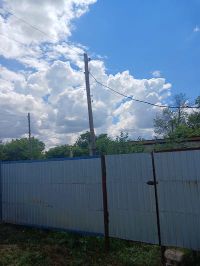 In the city of Жалтыр, Kazakhstan, an outdoor scene captures a cloudy sky with patches of blue and a large, white cumulus cloud formation. Two wooden utility poles with wires strung between them are visible. In the foreground, a corrugated metal fence runs horizontally across the frame, with a gate on the right side secured by a latch. Green foliage and grass are visible below and behind the fence, with some buildings and trees further in the background. The scene appears to be a residential or industrial area on a bright, partly cloudy day.