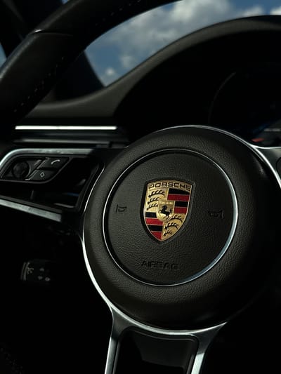 The image shows the interior of a Porsche vehicle, focusing on the steering wheel. The Porsche emblem is prominently displayed in the center of the steering wheel. Various buttons and controls are visible on the steering wheel's spokes. The surrounding interior is dark. The scene takes place in Diurtiuli, Russia.
