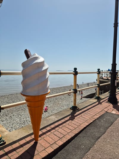 The image captures a sideways view in Penarth, United Kingdom, featuring an oversized ice cream cone sculpture positioned near a seaside promenade. The cone is primarily white with a brown chocolate flake. The promenade, lined with a red brick walkway and black asphalt strip, is bordered by a low wooden fence. The backdrop includes a pebble beach and a calm sea under a clear blue sky. Distant figures of people are scattered along the beach. A lamppost is visible in the top left corner of the frame.