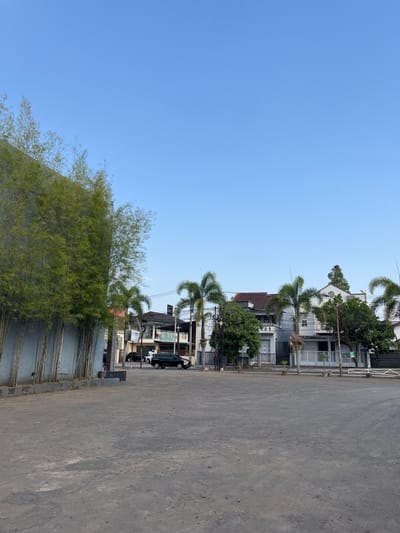 The image depicts an outdoor scene with a clear blue sky above. In the foreground, a large, flat expanse of grey, dusty ground dominates the frame. To the left, a tall, grey concrete wall is partially obscured by a dense cluster of tall, slender bamboo stalks with vibrant green leaves. Beyond the bamboo and wall, a street is visible with several buildings and parked cars.

The buildings appear to be residential or commercial, with multiple stories and varying architectural styles. Some have white facades, while others feature darker roofs and accents. Palm trees and other lush green foliage are interspersed among the buildings, adding a tropical feel to the environment.

On the street, a black SUV is parked, and a white car is also visible. The presence of power lines and poles suggests an urban or suburban setting. The overall impression is of a quiet, possibly late afternoon scene, with the sunlight casting soft shadows on the ground.

While the image does not provide explicit clues to its exact location within the city of Garut, Indonesia, the presence of palm trees, the architectural styles of the buildings, and the clear, bright sky are consistent with many areas in the region. The scene suggests a location that is developed but also retains a significant amount of natural greenery.