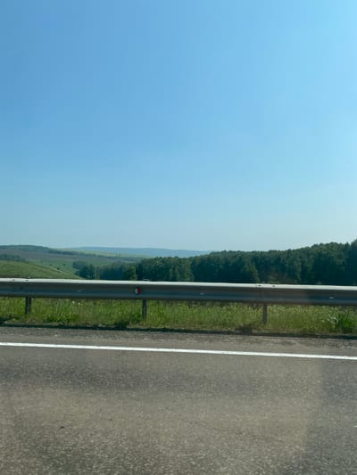 A wide-angle, slightly tilted, outdoor, eye-level shot captures a pastoral landscape under a clear, pale blue sky. In the foreground, a gray asphalt road with a white line marking its edge runs horizontally across the bottom of the frame. Beyond the road, a metal guardrail separates the pavement from a grassy verge dotted with wildflowers. The middle ground reveals rolling green hills and fields, with patches of darker green forest marking distant ridgelines. The overall impression is one of serene natural beauty, likely captured from a moving vehicle on a sunny day. The location is identified as Chulym, Russia.