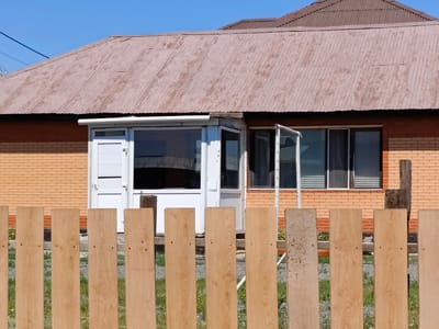 In the city of Қосшы, Kazakhstan, a low-angle shot captures a portion of a single-story brick house with a corrugated metal roof. The house is predominantly constructed of light orange brick, with a lighter orange brick base. A white enclosed porch with large windows is situated in the center of the visible facade. To the right of the porch, two windows with blinds are visible. The roof, made of weathered, reddish-brown corrugated metal, shows signs of age and rust. In the foreground, a light-colored wooden plank fence runs horizontally across the frame, obscuring the lower part of the house and the grassy ground beyond. The sky is a clear, bright blue.