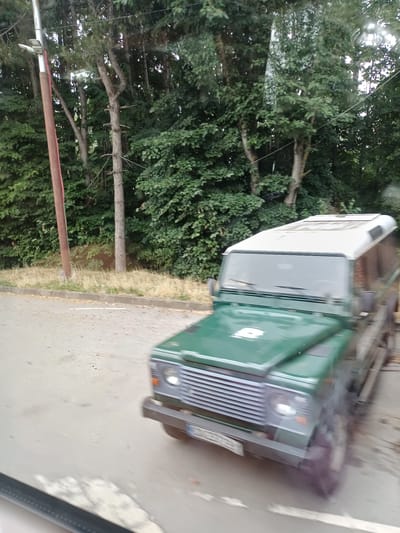 A green off-road vehicle with a white roof is captured in motion, blurred due to movement. The vehicle is positioned on a paved surface, possibly a road or parking area, adjacent to a grassy verge and a dense line of trees. A tall, reddish-brown pole with a light fixture is visible on the left side of the image, alongside the trees. The background is dominated by lush green foliage from the trees. The scene appears to be in a natural or semi-natural setting, with no specific identifiable urban features of Iovkovo, Bulgaria, directly discernible from the image alone, other than the implied location context.