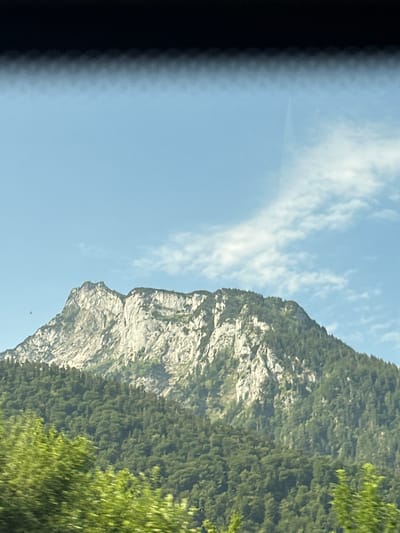 This image depicts a mountainous landscape with a prominent rock face covered in green vegetation. The sky is a clear blue with scattered white clouds. The scene appears to be captured from a moving vehicle, as indicated by a blurred dark area on the left side of the frame, suggesting motion. The sunlight illuminates the rock face, highlighting its rugged texture.

The orientation of the image is rotated, with the mountain rising from the bottom left and extending towards the top right. A noticeable feature is a formation on the rock face that resembles a human profile or a face, with a distinct nose and chin. The lower part of the mountain is densely covered with trees, creating a green, textured appearance. In the distance, further trees can be seen, also covered in green foliage. A small, dark object, possibly a bird or a drone, is visible in the sky near the mountain.

The time of day appears to be daytime, likely late morning or afternoon, given the bright sunlight. The weather seems pleasant and clear. The location is identified as Glanegg, Austria, which is consistent with a mountainous, forested environment. There is no visible text in the image. The overall mood is one of natural beauty and a sense of adventure, perhaps a scenic drive through the Austrian Alps.
