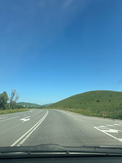 The image depicts a view from a vehicle traveling on a paved road in the city of Aya, Russia. The road has double yellow dividing lines and white lane markings, including directional arrows. To the left of the road, there is a grassy verge with trees, one of which is bare and appears dead. Beyond the verge, a line of green hills stretches into the distance, under a clear blue sky. To the right of the road, a large, grassy hill dominates the landscape, dotted with scattered trees. The weather appears to be sunny and clear. The foreground of the image shows the dashboard of the vehicle.