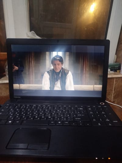 A Toshiba laptop is open and displaying a film. The film shows a man with dark, wavy hair wearing a white shirt and a dark vest with ornate detailing, and a dark scarf tied around his neck. He is looking to his right with a slight smile. In the background of the film, a person in a dark blue outfit is visible. The laptop is positioned on a wooden surface. The background of the room shows a window with light streaming in and a shelf with various objects, including a green container and a white bottle. The scene is indoors.