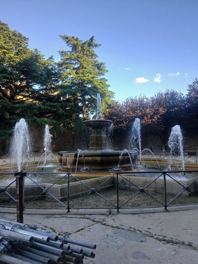 A multi-tiered fountain is in operation in what appears to be a city park or plaza. The fountain features several spouts of water cascading down stone levels and spraying outwards. The fountain is enclosed by a low, dark metal fence with a criss-cross pattern. Surrounding the fountain is a paved area with some patches of dry grass within the fenced enclosure. In the background, there are large, mature trees with dark foliage, some of which appear to be coniferous. Behind the trees, a stone wall is visible. The sky is a clear blue with a few wispy white clouds. In the foreground, a stack of silver metal pipes, possibly scaffolding or construction materials, lies on the ground. The scene is bathed in sunlight, suggesting it is daytime. The location is identified as Ávila, Spain. There are no people clearly visible in the frame.