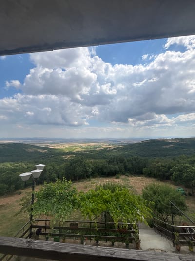 This is a vertical, medium-shot, eye-level outdoor photograph taken from a high vantage point. The scene depicts a scenic landscape with a vast expanse of rolling hills and a distant flat plain under a cloudy sky. In the foreground, a dark wooden railing runs along the bottom edge of the frame. To the left, a lamp post with three lights extends upwards. Green trees and foliage dominate the mid-ground, with a pathway leading downwards through them. The background reveals a wide, flat landscape with fields and distant hazy hills. The sky is filled with a mix of white and grey clouds against a bright blue backdrop. The overall impression is a tranquil and expansive view of nature. The location is identified as Pobeda, Bulgaria.