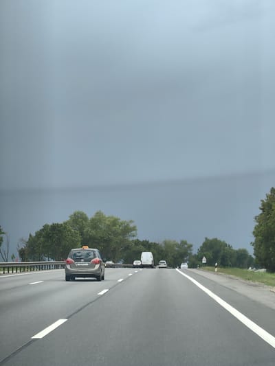 The image shows a highway scene with a grey sky overhead. In the foreground, a grey minivan with a yellow taxi sign on its roof is driving on the right side of a multi-lane asphalt highway. The vehicle has a Belarusian license plate that reads "3523 CT-7". White lane markings are visible on the road. To the left of the highway, a metal guardrail runs alongside a strip of grass. Beyond the guardrail, a line of green trees with dense foliage creates a backdrop. In the distance, other vehicles, including a white van and several cars, are also on the highway. The overall environment suggests an overcast day on a highway in Belarus.
