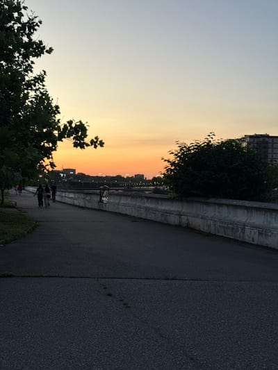 The image depicts a sunset scene along a paved walkway next to a body of water, possibly a river. The sky transitions from a light blue to a warm orange glow near the horizon, indicating the time of day is late evening. In the foreground, silhouetted trees with lush green leaves frame the left side of the image.
Several individuals are visible on the walkway, appearing to stroll or walk. Their figures are dark against the lighter background, suggesting they are facing away from the setting sun. The walkway itself is dark asphalt, with a concrete barrier running alongside it, separating it from what appears to be a body of water or embankment. Beyond the barrier, faint lights suggest buildings or a distant cityscape.
The overall mood of the image is tranquil and serene, characteristic of an evening walk during good weather. There is no visible text in the image. The location is identified as Khachta, Russia.