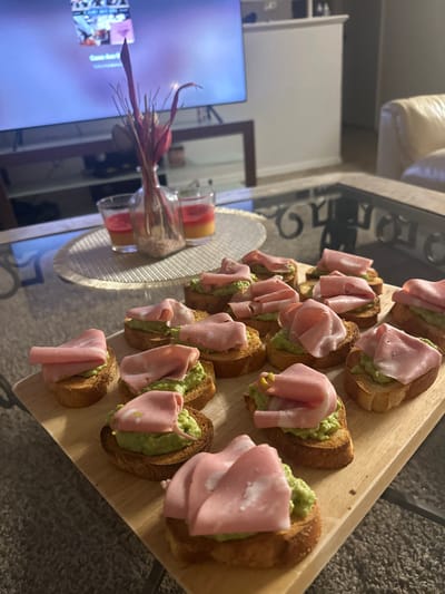 This image depicts an indoor domestic setting, likely a living room, within East Naples, United States of America. The primary subject is a wooden rectangular serving board positioned on a glass-top coffee table, displaying approximately 18-20 individual appetizers. Each appetizer consists of a toasted bread crostini topped with a green, mashed substance, likely avocado or guacamole, and a folded slice of light pink deli meat, consistent with mortadella.
To the left of the serving board, on the glass table and atop a circular light grey placemat, are three decorative items: a clear glass bottle vase containing dried reddish-purple foliage, flanked by two clear glass votive candle holders. The candles contain layered liquids, one red and the other orange/yellow.
In the blurred background, a large flat-screen television is visible, displaying indistinct content with a purple and white color scheme. The television rests on a dark brown rectangular console table. A light-colored, possibly cream or white, upholstered furniture piece, consistent with a sofa or armchair, is partially visible in the far right background. The floor is covered with light-colored carpeting. No individuals are visible in the scene. The lighting suggests an interior, possibly evening, ambiance.