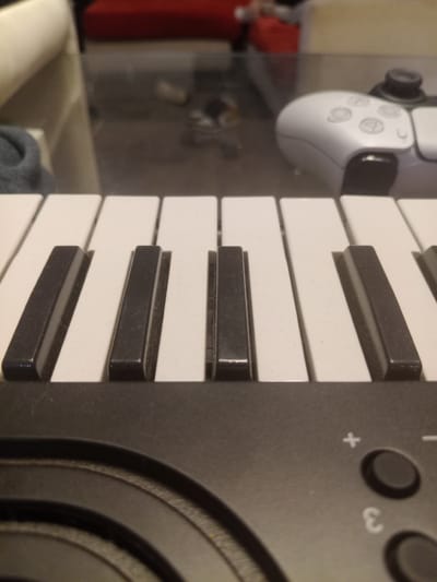 The image shows a close-up view of a keyboard, with a PlayStation 5 controller visible in the blurry background. The keyboard features white and black keys and buttons with markings like "+", "-" and "3". The controller is white. The background is out of focus, showing a sink and other miscellaneous objects. The image location is unknown; there's no information in the provided image that implies Lomas de Zamora, Argentina.