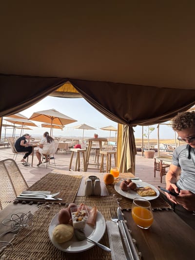 The image depicts an outdoor dining area, likely a restaurant or cafe, set up under a large tent in Agafay, Morocco. The scene is bathed in warm, natural light, suggesting it is daytime. In the foreground, a table is laid out for breakfast, featuring two plates of food. One plate contains a portion of yogurt with fruit, a bread roll, two hard-boiled eggs, and slices of ham. The other plate holds three hard-boiled eggs and a piece of toast. A glass of orange juice sits beside the latter, with an orange also visible on the table. Cutlery, napkins, and salt and pepper shakers are also present.

In the midground, two women are seated at separate tables, engaged in conversation. They are both casually dressed and appear to be enjoying their time. Beyond them, the dining area extends with more tables and chairs, many covered by large, beige umbrellas, indicating an effort to provide shade. In the background, the landscape of Agafay unfolds, characterized by arid, rolling hills and a clear, bright sky. The overall atmosphere is one of relaxed outdoor dining in a desert environment.
