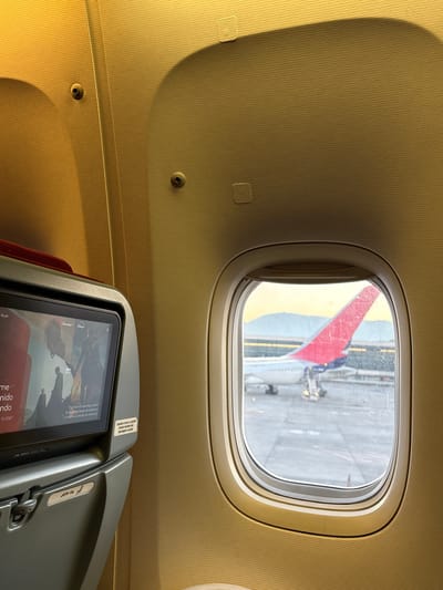 A passenger's view from inside an airplane, possibly at Pudahuel, Chile, given the language on the entertainment screen. The window offers a view of the tarmac and part of the airplane's red and yellow wing. An in-flight entertainment screen is visible in the foreground.
