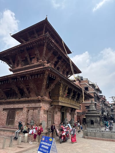 A medium, long-shot image depicts the Patan Museum in Lalitpur, Nepal. The scene includes a multi-tiered, brick-and-wood temple, elaborately carved and decorated. People are sitting and standing around the base of the temple, some on steps, others near a bell. A sign indicates the "Lalitpur Metropolitan City and Patan Museum Tourist Entrance Fee." A scooter is parked nearby. Overhead, the sky is partly cloudy. The architecture and scene suggest a historical and cultural site.