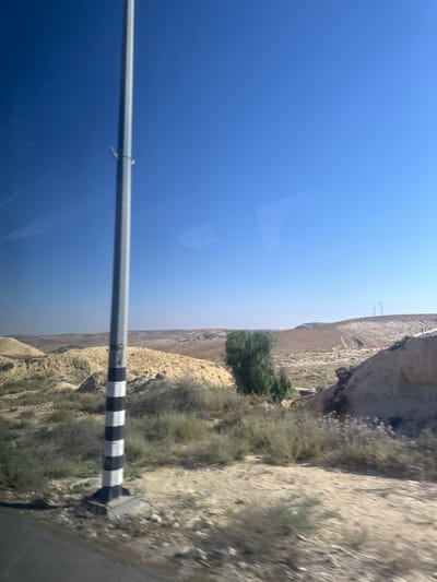 The image depicts a roadside scene, likely captured from a moving vehicle, given the slight blur and the perspective. The dominant elements are a vast, clear blue sky that takes up a significant portion of the frame and a rugged, arid landscape to the right. The landscape features dry, rocky terrain with sparse vegetation, suggesting a desert or semi-arid environment.
In the foreground, a silver metal pole runs horizontally across the lower part of the image. It has a band of black and white stripes near its right end, which appears to be part of a marker or warning sign. The pole is attached to something out of frame to the left, and to a rectangular concrete base on the right.
The background reveals more of the dry landscape, with hills and valleys. A few small trees or bushes dot the terrain, and a single, larger, bushy green tree is visible further back, providing a contrast to the otherwise muted earth tones. In the far distance, some structures, possibly a small town or scattered buildings, can be seen on the horizon. Two simple poles that resemble utility poles or light posts are also visible on a distant hillside.
The bright, cloudless sky indicates a sunny day, likely during daylight hours. The overall impression is one of a vast, open, and dry natural environment. No people or specific activities are depicted, other than the implied travel. The location context provided points to Arad, Israel, which aligns with the arid landscape. There is no discernible text in the image.