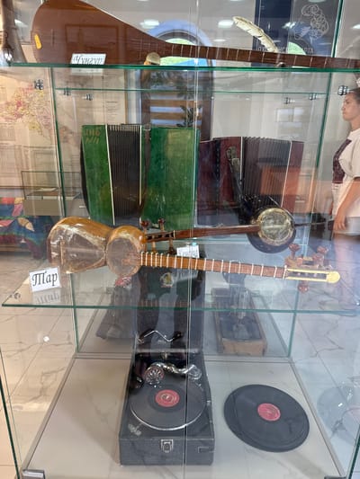 The image shows a museum display case filled with musical instruments and related items. In the top shelf, a long-necked stringed instrument is visible, along with a label. The second shelf holds two accordions, one green and one dark red, and another stringed instrument with a round body. A label is attached to this instrument. The bottom shelf features an old-fashioned record player with two vinyl records. The museum appears to be located in Khuchni, Russia, as suggested by a map visible in the background of the second shelf, which is labeled with the city name. A person is partially visible on the right side of the image, looking into the display case. The environment is indoors, with the museum exhibits presented behind glass.