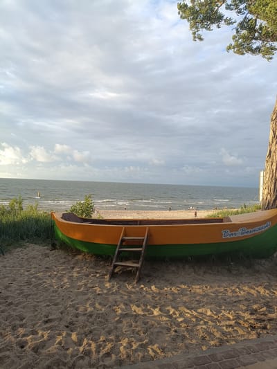 This media file depicts a serene beach scene in Stegna, Poland. In the foreground, a vibrant orange and green boat rests on the sandy shore. A wooden ladder leans against the boat, suggesting a connection to the water or perhaps an artistic display. The boat bears visible text that reads "Bar Bosmank".
Beyond the boat, the beach stretches towards the sea, where gentle waves roll onto the shore. Several people can be seen in the distance, enjoying the beach and the water. The sea is a deep blue-green, meeting a cloudy sky with patches of lighter grey and white. Tall green grass grows along the edge of the beach, adding to the natural beauty of the setting. To the right of the frame, the rough bark of a large tree trunk is visible, its branches reaching out towards the sky.
The lighting suggests it is either early morning or late afternoon, with soft sunlight casting shadows on the sand. The overall mood is peaceful and tranquil, evoking a sense of a relaxed day at the seaside. The combination of the boat, the sea, the sand, and the distant figures creates a classic beach atmosphere.