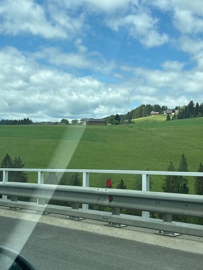 The image shows a view from a vehicle traveling on a road in Unterauerling, Austria. A metal guardrail is visible on the right side of the road, partially obscuring the view. Beyond the guardrail, there is a green field with trees along the edge. In the distance, there are buildings that look like rural Austrian houses. The sky is blue with white clouds. The photo is taken at a slight angle, and it is bright and sunny.
