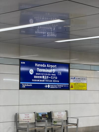 The image shows a subway or train station interior with signage and seating. A large blue sign displays information about "Haneda Airport Terminal 3" in English, Japanese, and Korean, with a "KEIKYU" logo at the top left. The sign indicates connections to "Tenkubashi" station, and also mentions "Haneda Airport Terminal 1.2". To the right of the blue sign is a yellow information placard from ANA (All Nippon Airways) regarding international flights to terminals 2 and 3. In the foreground, there are two metallic benches with signs on them displaying "Welcome" in multiple languages including English, Korean, and German. The environment appears to be modern and clean, with white tiled walls and illuminated ceiling panels. The lighting is bright, likely from overhead fixtures. The scene depicts an indoor transportation hub, likely within Ota, Japan, given the explicit mention of Haneda Airport. There are no people visible in the image.