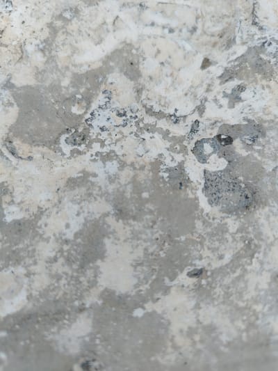 The image shows a close-up, slightly blurred view of a concrete surface. The concrete exhibits a rough texture with variations in color, including shades of gray, white, and some darker speckled areas. There are patches of what appears to be chipped or worn material, creating an uneven and aged appearance. The lighting suggests a diffuse light source, casting minimal shadows. The context of Stepnoe, Russia is not visually discernible from the image itself.