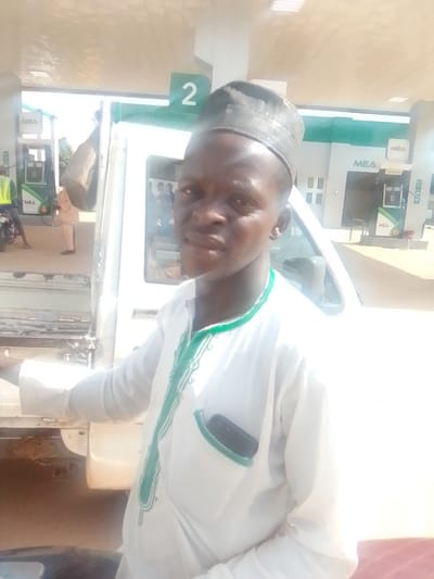 A young man wearing a white robe with green trim and a dark patterned cap stands in front of a white vehicle at a gas station. He is looking towards the camera, and his face is partially obscured by sunlight. A cell phone is visible in his robe's pocket. In the background, there are gas pumps with "MEA" signage, and another person in a neon vest is standing near a motorcycle. The location is the BYE PASS gas station in Nigeria. The overall scene is brightly lit by natural sunlight.