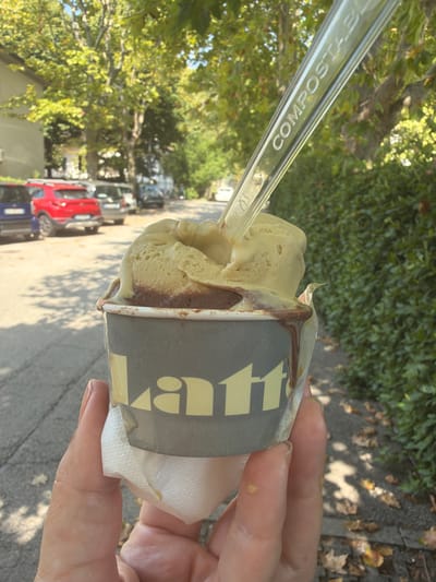 The image shows a close-up of a person holding a cup of gelato. The gelato is a light brown and green color and is melting down the sides of the cup. The cup has a logo that says "Latt" in yellow letters. A clear plastic spoon is stuck in the gelato. In the background, there are trees, parked cars, and a sidewalk. The lighting suggests it is daytime. The spoon has the word "COMPOSTABLE" embossed on it.