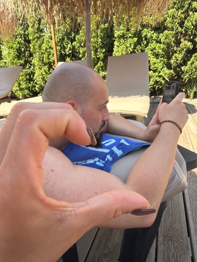 The image shows a man lying down, possibly on a lounge chair, outdoors. He is holding a smartphone in his right hand and appears to be looking at it. A blue towel is visible on his lap. A person's hand, in the foreground, is positioned to appear as if it is interacting with the man or the phone. The background includes a thatched umbrella, lush green bushes, and wooden decking. The scene suggests a relaxed outdoor setting, potentially a resort or a backyard with leisure furniture. There is no information in the image to suggest it is located in Plovdiv, Bulgaria.