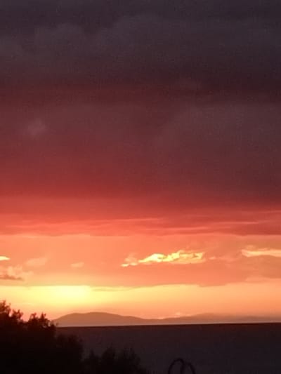 The image shows a sunset scene in Pyli, Greece. A portion of a tree is visible in the lower right corner. The sky transitions from dark on the left to orange on the right, where the sun is setting behind clouds.