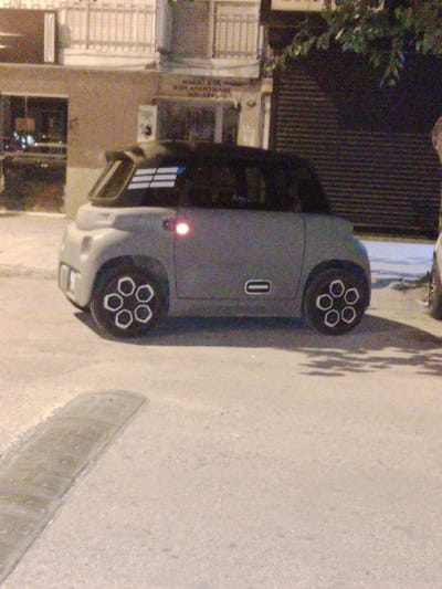 The image shows a small, grey, futuristic-looking car parked on a paved street at night. The car has a distinctive design with large, circular wheel covers that have a flower-like pattern. A red light is visible on the side of the car, possibly a taillight or indicator. The background shows a building with a dark, corrugated metal shutter and some greenery above it. Part of a building with a balcony and railings is visible to the left. The overall lighting suggests it is nighttime, and the shadows on the ground indicate streetlights or lights from the building. The scene appears to be in an urban environment. The car itself is the primary subject, and its unique design is a notable detail. There is some faint text on the building in the background, which appears to read "MAHAL KOR" and "KOR APARTMANI", but it is largely illegible due to the image quality and distance. The location context provided is Karşıyaka, Turkey. No people are visible in the image.