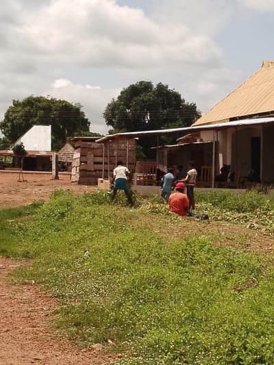 In Adikpo, Nigeria, a group of individuals is engaged in work near a cluster of buildings under a cloudy sky. In the foreground, a person wearing a red t-shirt and dark trousers sits on the ground amidst green vegetation. To their left, another person in a light blue shirt and dark trousers stands. To the right of the seated individual, a person in a white t-shirt and dark trousers is observed from behind. Further to the right, another person is partially visible. In the background, to the left, a white metal-roofed structure stands, and next to it, a building with a light-colored, corrugated metal roof extends towards the right. A stack of wooden materials is positioned in front of a building with a covered porch supported by poles. Chairs and tables are arranged under the porch. The ground is a mixture of dirt and grass.