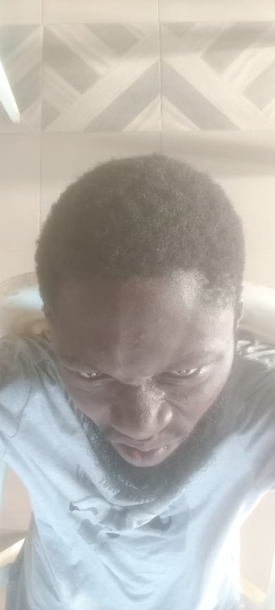 A low-angle selfie depicts a dark-skinned man with short, dark hair and a beard. He is wearing a light-colored t-shirt. The background consists of tiled walls with a geometric pattern. The location is indeterminate, and the connection to Bende, Nigeria, is unsubstantiated from the visual data.