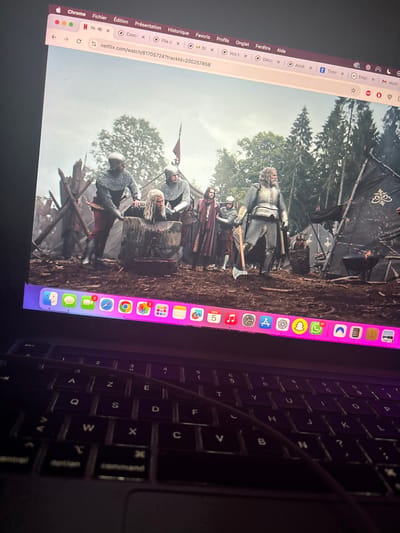 A MacBook laptop, physically located in Maisons-Alfort, France, displays a full-screen video from Netflix via the Chrome browser. The macOS menu bar is visible at the top, showing standard application menus and the URL `netflix.com/watch/81705724?trackid=200257858`.
The video content depicts an outdoor, muddy scene in what appears to be a medieval or fantasy encampment, flanked by evergreen trees and tents. In the central foreground, two men in chainmail armor and helmets are physically restraining another man with long, light-colored hair over a large, crude wooden block or stump, suggesting an imminent beheading or execution. Several other armed men in period attire, including one prominent figure clad in heavy chainmail holding a battle-axe, stand observing the scene to the right. The background features more armed figures, wooden barricades, and a dark tent with a decorative gold emblem, along with a small fire or brazier.
Below the video, the macOS Dock is visible, illuminated with a slight pinkish hue, displaying numerous application icons. Several icons show notification badges, including FaceTime (2), Calculator (2), Calendar (5), Music (1), and WhatsApp (1). The date "NOV 5" is visible near the Calendar icon. The laptop's keyboard, featuring an AZERTY layout and faint purple backlighting, occupies the lower portion of the image. A dark cable runs diagonally across the keyboard from the top-left to the bottom-right. The ambient lighting suggests a dim environment around the laptop.