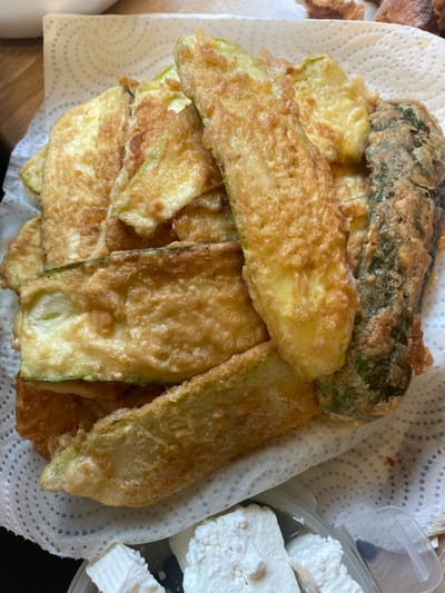 A pile of golden-brown fried zucchini slices rests on a white paper towel, which is dotted with blue patterns. Some slices are whole and elongated, while others appear to be broken or overlapping. The zucchini has a crispy, battered coating, and some pieces show a hint of green from the skin. In the lower right corner, a clear plastic container holds several rectangular pieces of white cheese, possibly feta. The background is blurred, showing a wooden surface and a hint of other food items. This scene is depicted in Gerstheim, France.