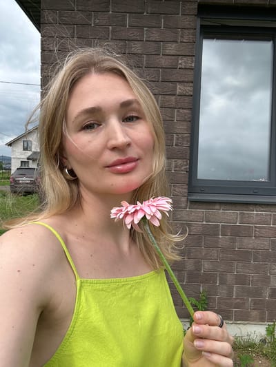 A young blonde woman holds a pink gerbera daisy in front of her face, with the flower obscuring part of her lips and chin. She is wearing a neon green sleeveless top. She has on a silver hoop earring in her left ear and a dark ring on her right ring finger. She is standing outdoors in front of a brick building with a dark-framed window. In the background, a white house with a dark roof and a dark car are visible. The scene appears to be in a rural or suburban area, possibly the city of Mokino, Russia, given the architectural style and vegetation. The sky is overcast.