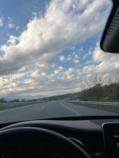 The image depicts a view from inside a car on a highway near Valkovo, Bulgaria. The sky is filled with clouds, some white and fluffy, with patches of blue sky visible. The highway stretches into the distance, flanked by trees and guardrails. A vehicle dashboard and steering wheel are visible in the foreground.