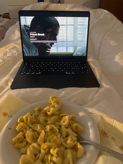 This image, taken in Basiglio, Italy, captures a cozy indoor scene from an overhead perspective, centered on a laptop and a plate of food resting on a bed. A black laptop, likely a MacBook, is open on a white bedspread, displaying a scene from the television show "Prison Break." The laptop screen shows a close-up of a man's face, appearing to be of African descent, with short dark hair, looking towards the top of the frame. Vertical blinds are visible in the background through what appears to be a window. Text overlaid on the screen identifies the show as "Prison Break Season 1, Bluff: Ep. 18." Subtitles read, ""Invisible handcuffs." Handcuffs on your mind." A "Paused" icon is faintly visible at the top right of the screen.

To the right of the laptop, a white, round plate is filled with a generous serving of tortellini or a similar pasta, covered in a light sauce. A silver fork is resting in the pasta. The bed is made with white bedding, and a light-colored blanket or throw, patterned with what appear to be yellow sunflowers, is peeking out from beneath the plate. The scene suggests a relaxed leisure activity, likely in the evening, involving watching a show and enjoying a meal in bed. The lighting is soft, hinting at an indoor setting with some ambient light, creating a comfortable atmosphere.