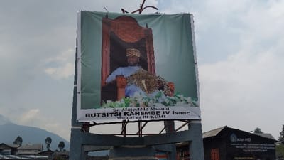 A large banner shows a portrait of Mwami Butsitsi Kahembe IV Isaac, the Mwami de Bukumu. He is seated on a throne, wearing a leopard-print headdress and shawl over light blue clothing. Below the portrait, the banner identifies him by name and title. Behind the banner are utility poles or antennas. To the right is a building with the acronym "ACAV-MID" and some French text. The background includes trees, other buildings, and a mountain range. The sky is cloudy.