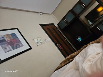 The image is taken inside a hotel room in Paranaque City, Philippines. The room is in disarray with a bed in the foreground, an open door on the left side, and an open closet on the right side. The bed has a white sheet and a brown blanket on it. The closet has shelves with various items on them.  There are a few items in the background on the top shelf of the closet including a clear bottle of water, a blue trash can, and a light shining in the distance.  The door is partially open and appears to be closed to a hallway.  The wall is beige and has a framed picture on the left side and a sign on the right side.  There is a white light switch on the wall near the open door.  The ceiling is white.  The image was taken at latitude 14.521 and longitude 120.994.  The image appears to be taken from the floor looking up towards the ceiling.  The image is slightly out of focus.  The image is taken from the perspective of someone lying in the bed.