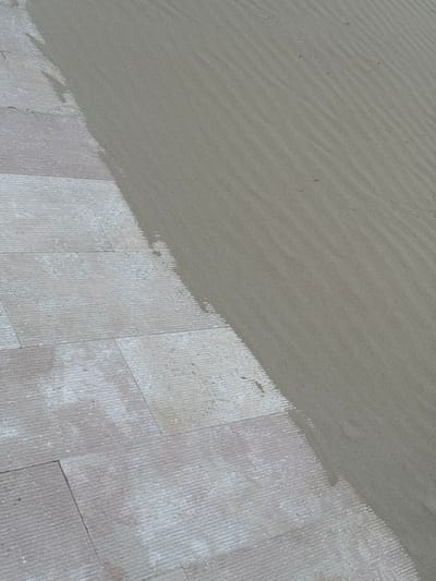 The image displays an outdoor ground-level scene in Grottammare, Italy, depicting the transition between a paved surface and a sandy area. The lower-left portion features light-colored, rectangular paving stones with visible parallel striations on their surface, possibly indicating a textured finish or wear. These stones are arranged in a regular pattern, and some exhibit areas of lighter discoloration. The upper-right portion consists of fine-grained, light brown-gray sand. The sand displays distinct, parallel ripple patterns, oriented diagonally across the frame, consistent with wind or water movement. Scattered small, dark specks of debris are visible within the sand. An irregular, undulating edge delineates the boundary, with sand partially encroaching onto the paved surface. No people, animals, or other significant objects are present in the scene.
