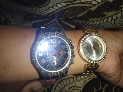 A close-up image displays a human arm, with medium brown skin tone, wearing two wristwatches simultaneously. On the left is a large, dark-colored "KNUODE K1720" branded chronograph-style watch with a dark dial, visible subdials, red accents, and a tachymeter scale on its bezel. To its right, a smaller, gold-toned "GUCCI" branded watch features a round, crystal-encrusted bezel, a silver-toned textured dial with the brand logo, and a metallic link bracelet. The watches are positioned side-by-side on the wrist, with a hand partially visible at the bottom of the frame. The background consists of a dark fabric with a light-colored ornamental pattern. The image contains no visual information to determine its specific location within the Umsobomvu Local Municipality, South Africa.